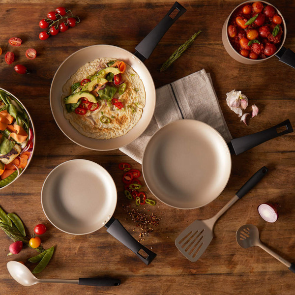 Set of beige non-stick frying pans and utensils arranged on wooden table with fresh vegetables