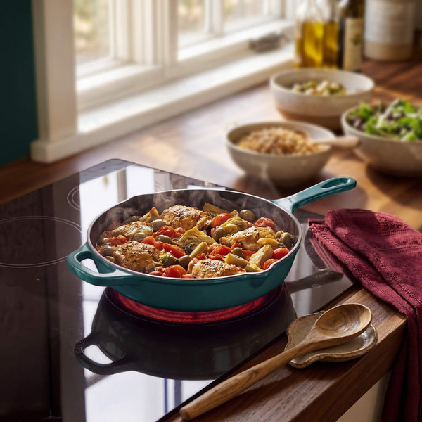 Green cast iron frying pan with cooked chicken and vegetables on a hob