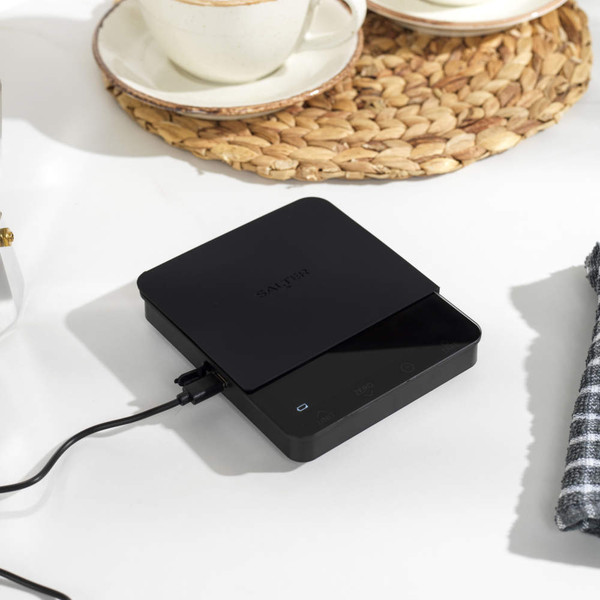 Black digital kitchen scales with slim platform, shown plugged in on a worktop