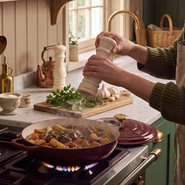 Cream pepper grinder seasoning stew in burgundy cast iron casserole on gas hob