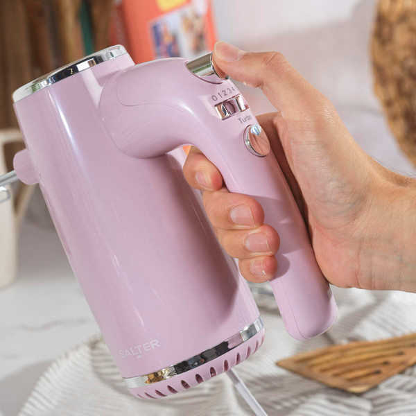 Pink hand mixer in use, shown close-up with speed controls and beaters visible.