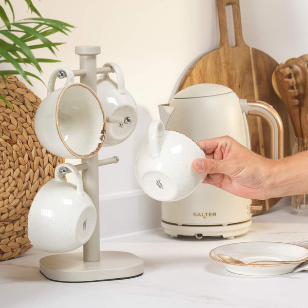 Set of four cream speckled stoneware mugs on stand, hand holding a mug in kitchen setting