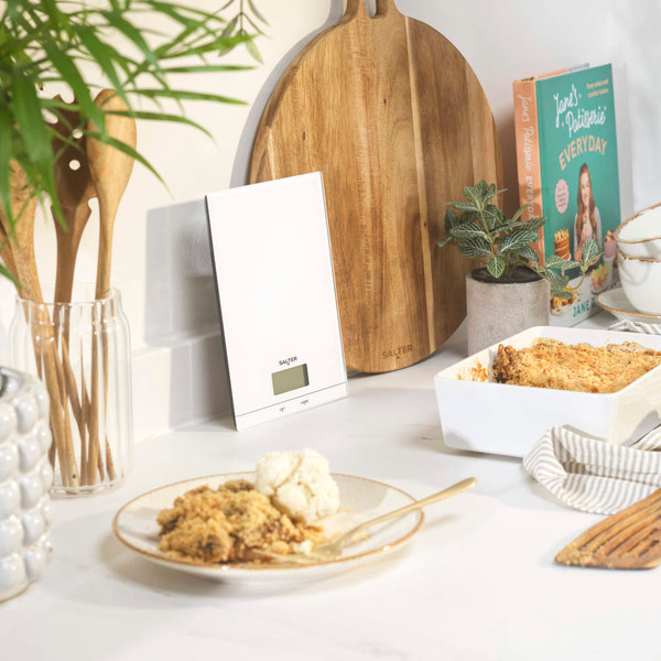 White digital kitchen scale on countertop beside wooden utensils and baking dishes.