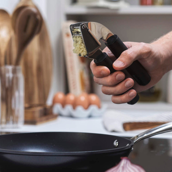 Hand using black garlic press over frying pan in kitchen