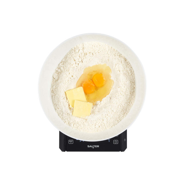 Baking ingredients in white bowl on Salter kitchen scale: flour, butter, and eggs.