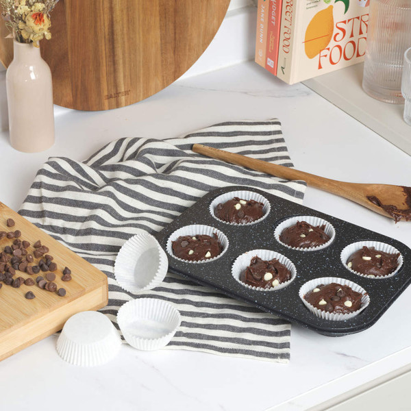 Six chocolate chip muffin batter portions in white paper cases in a non-stick tray on kitchen worktop.