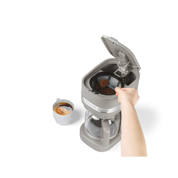Hand adding coffee grounds to open taupe filter coffee maker, with cup of brewed coffee nearby