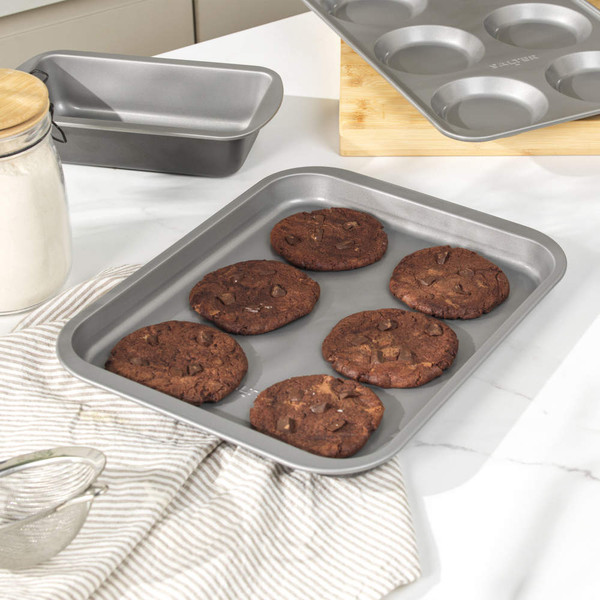 Baking tray with six chocolate cookies, loaf tin and muffin tray on kitchen worktop.