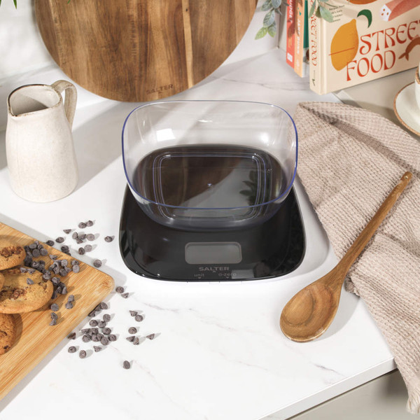 Digital kitchen scale with clear bowl on countertop, cookies and utensils beside.