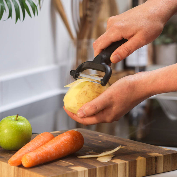 Black-handled peeler in use on potato, carrots and green apple on chopping board.