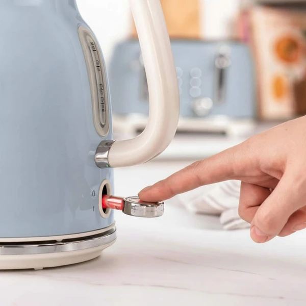 Blue electric kettle with cream handle, power switch being pressed on a kitchen counter.