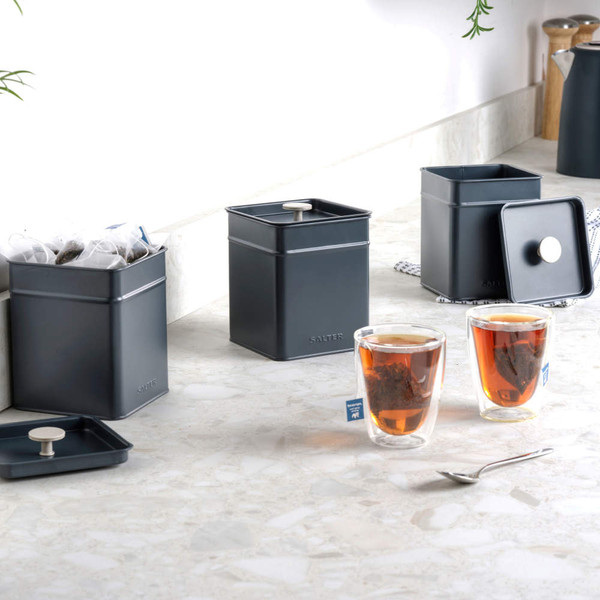 Set of three matte black square storage tins with lids, shown on a kitchen worktop with tea bags and tea glasses.