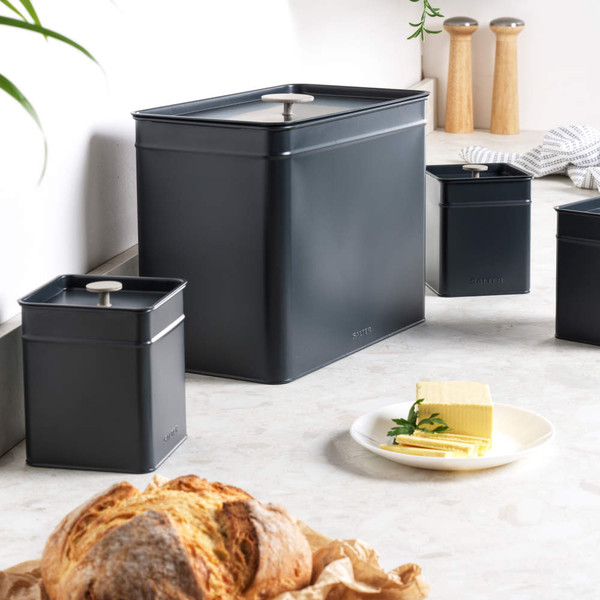 Set of grey metal kitchen storage canisters on countertop, including bread bin, with loaf and butter.