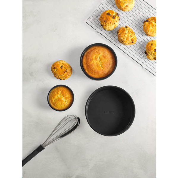 Round black baking tins with cakes and scones, plus a whisk, shown on a light background