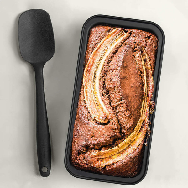 Non-stick black loaf tin with freshly baked banana bread and a black spatula beside it