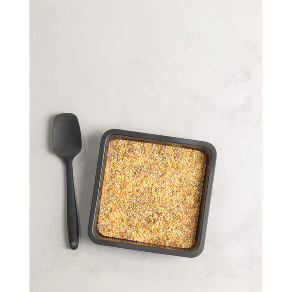 Square black tin filled with golden crumble topping beside a black spatula, shown overhead.