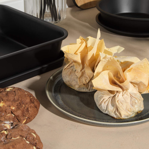 Black non-stick roasting tin and round baking tins with cookies and filo pastry parcels