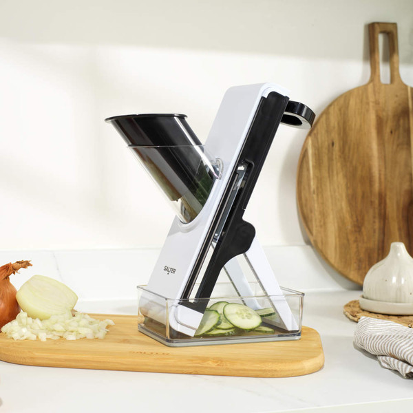 White and black kitchen slicer cutting cucumber on chopping board, onion and tomato beside