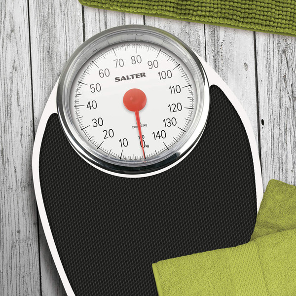 Black mechanical bathroom scale with large dial, shown on wood floor with green towels.