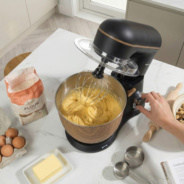 Black and wood stand mixer blending cake batter on kitchen counter with baking ingredients.