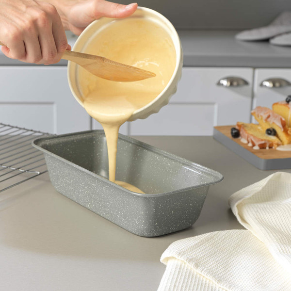 Cake mixture being poured into a grey non-stick loaf tin on kitchen countertop