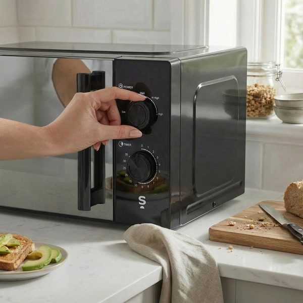 Person adjusting black microwave dials on kitchen counter, loaf and avocado toast in view