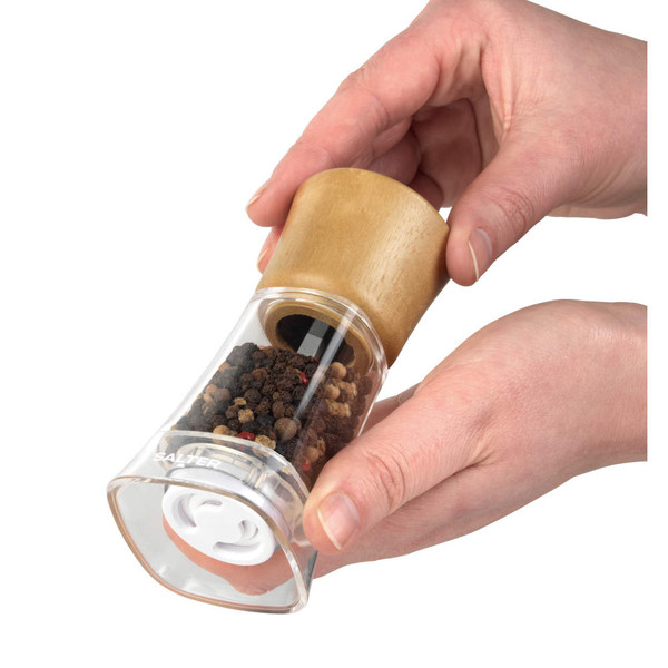 Manual pepper mill with clear body and wooden top, held in both hands and filled with peppercorns