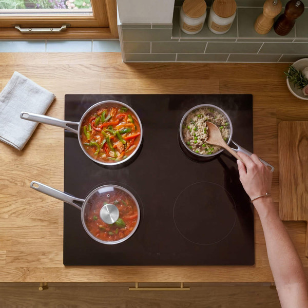 Overhead view of three silver saucepans cooking food on black induction hob in modern kitchen
