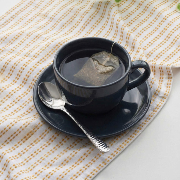 Black ceramic cup and saucer with tea bag and spoon on patterned cloth.