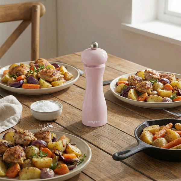 Pink manual pepper mill on table with roast chicken and vegetables, dining setting.