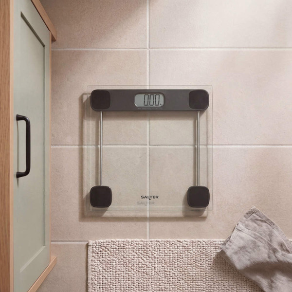 Digital bathroom scale with clear glass platform, black accents, displayed on tiled floor