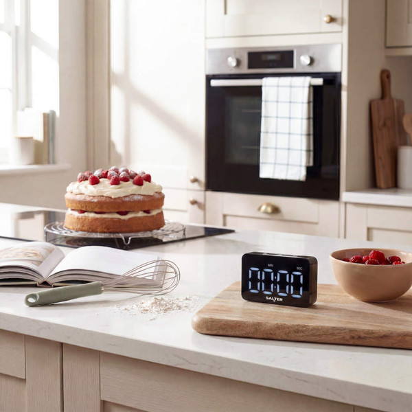 Digital kitchen timer on chopping board with baking ingredients and cake in modern kitchen