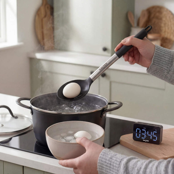 Salter kitchen timer beside boiling eggs in pot and slotted spoon with egg.