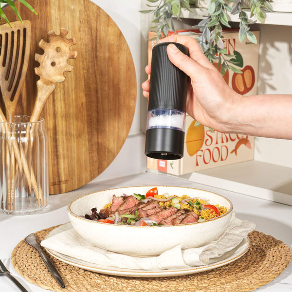 Hand using black electric salt grinder above bowl of steak salad on kitchen worktop.