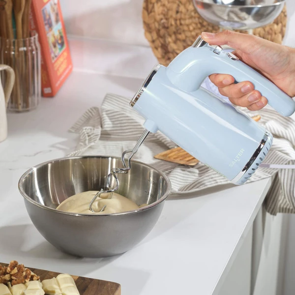 Hand mixing dough with a blue electric hand mixer in a metal bowl on a kitchen counter