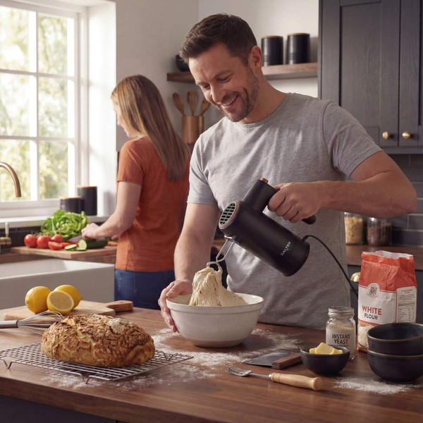 Person using black electric hand mixer with dough in kitchen, flour and yeast visible