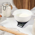 Digital kitchen scale with white mixing bowl on worktop, surrounded by baking ingredients