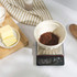 Cocoa powder weighed in a white bowl on digital kitchen scales, butter and flour beside on board.