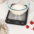 Glass bowl with milk being poured, on digital kitchen scale showing zero.