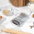 Digital kitchen scale with large clear mixing bowl on white worktop, shown with baking ingredients.