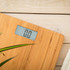 Digital bamboo bathroom scale with LCD display, shown on wooden floor beside white towel.