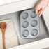 Grey six-cup muffin tray with speckled finish, shown being placed in kitchen drawer