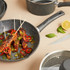 Grey non-stick frying pan with black handle, shown in use with food skewers; part of cookware set.