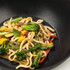 Vegetable stir fry with udon noodles in black wok, featuring broccoli and peppers