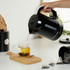 Black electric kettle pouring hot water into a glass mug with tea on a kitchen counter