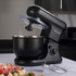 Black stand mixer on kitchen counter with cake, eggs, and baking tools.
