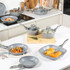 Set of grey non-stick pots and pans with glass lids, shown with cooked food in a modern kitchen