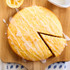 Round lemon drizzle cake with icing, one slice cut, on wooden board