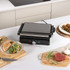 Black panini press with two toasted sandwiches inside, showing filling of ham, cheese and salad.