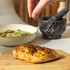 Hand sprinkling spices over cooked chicken breast on chopping board, with salad and mortar behind